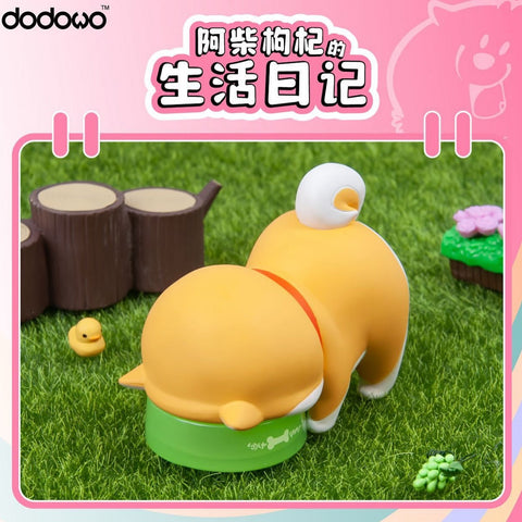 A Shiba Inu figure with its head in a green food bowl, backside up, tail curled. The playful pose is set on green grass with tree stump props and a small yellow duck nearby.