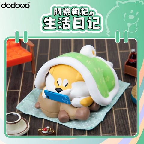A Shiba Inu under a green patterned blanket, holding a blue game console. The cozy pup lies on a mat with coffee, cake, and soda props scattered in the room.