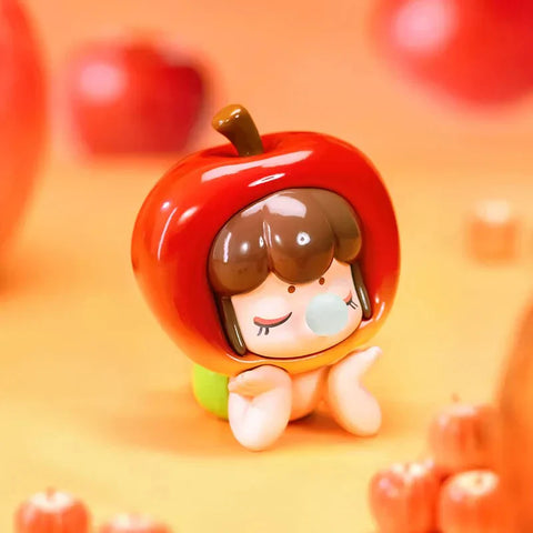 This image features a close-up of an adorable collectible figurine from the Baby Nanci Fruits Series Blind Box collection. The figure is designed with a peaceful expression, closed eyes, and small hands resting on its cheeks. It wears a glossy, vibrant red apple-shaped headpiece with a brown stem on top, resembling a fresh apple. The character has short brown hair with bangs, adding to its cute aesthetic. The warm orange-toned background complements the figure.