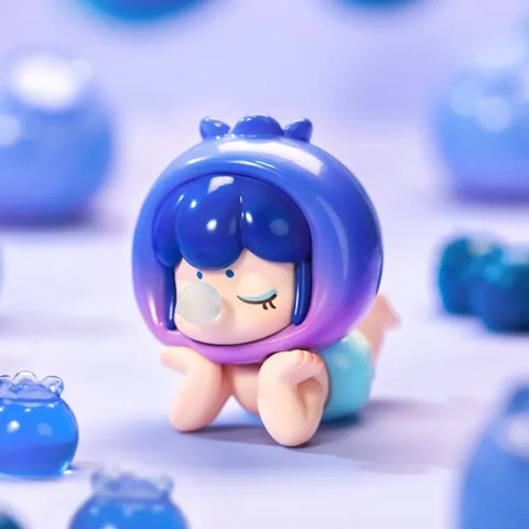 This image showcases an adorable collectible figurine from the Baby Nanci Fruits Series Blind Box collection. The figure has a calm, dreamy expression with closed eyes and hands resting on its cheeks. It wears a glossy, deep blue and purple blueberry-inspired headpiece, complete with small details resembling the crown of a blueberry. The character’s short, wavy blue hair complements the theme. The soft lavender background is adorned with various blueberry-shaped decorations.