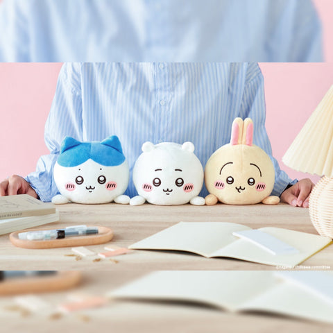 Three round plush characters—one blue-eared, one white, and one beige with rabbit ears—sit in a row on a wooden desk in front of a person wearing a blue striped shirt. Each plush has embroidered eyes, small paws, and pink blush. Notebooks, pens, and a lamp decorate the pastel-themed workspace.