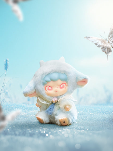 Seated baby‑lamb plush in pale blue fur with tiny white horns and a sky‑blue tassel charm rests in sparkly frost. Butterflies blur at the edges while a bright, cloudless sky adds crisp light to the character’s coral eyes and soft woolly details.