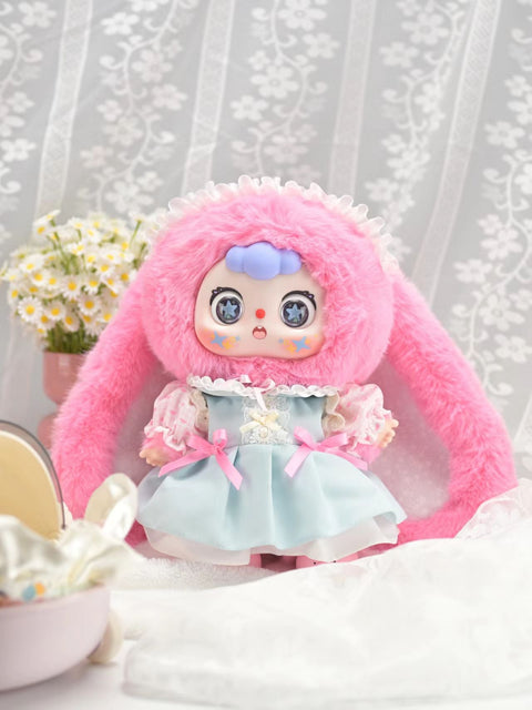 Doubao Frosting Rabbit appears in a pink plush bunny suit with long floppy ears and a pastel blue frilly dress tied with pink bows. Its big starry eyes, delicate lace details, and soft fuzzy textures enhance the whimsical design, posed sweetly in a light floral indoor setting.