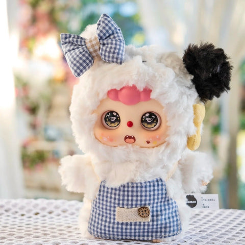 Puppy Doubao plush in a fluffy white suit with black ear accents and a blue gingham bow bib. Its expressive round eyes sparkle with puppy icons, and blushed cheeks enhance its mischievous but lovable look, making it a classic favorite from the plush blind box lineup.