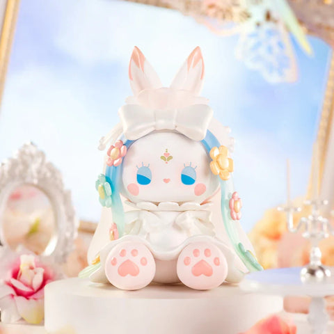 A delicate figurine from the "Emma Secret Forest: Beloved Series" sits in a dreamy, pastel setting. The character has rabbit-like ears, flowing blue and green hair adorned with small flowers, and a serene expression. It wears a white outfit with a large bow and soft pink paw pads. The ethereal background features ornate frames, flowers, and candles, creating an elegant, fairytale atmosphere that enhances the figure’s whimsical and graceful charm.