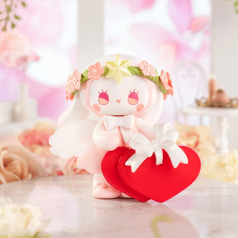 A sweet figurine from the "Emma Secret Forest: Beloved Series" radiates charm in a romantic, pastel setting. The character wears a soft pink dress, translucent wings, and a floral crown with a golden star. Holding a bright red heart-shaped box wrapped in a white ribbon, it symbolizes love and devotion. The background, featuring delicate roses, elegant décor, and warm pink tones, enhances the dreamy, fairytale-like atmosphere of this angelic and affectionate figure.