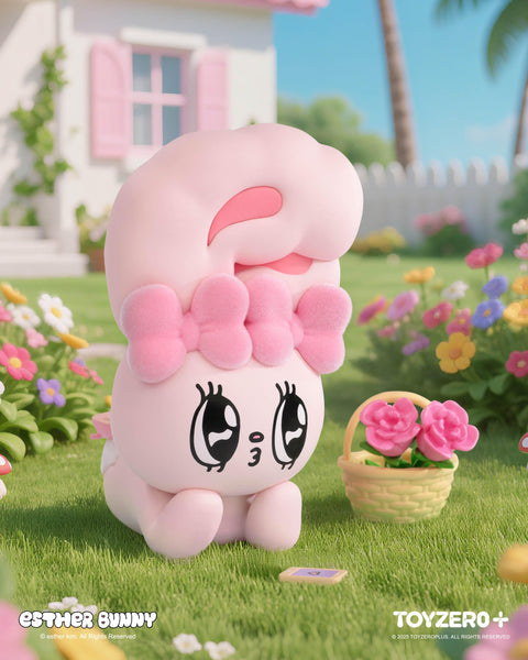 A pastel pink Esther Bunny figurine with large sparkling eyes and a fluffy bow lays adorably on the grass. Next to it is a small basket of pink flowers, surrounded by a garden of colorful blooms, with a white picket fence and cottage-style house in the sunny background.