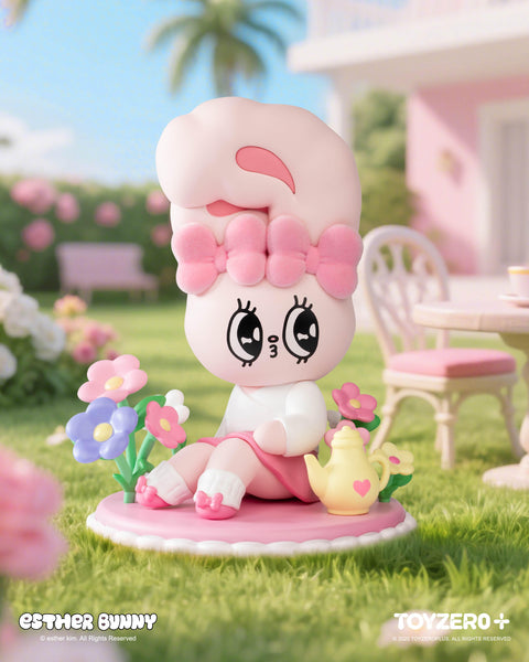 A pastel pink Esther Bunny figurine with a fluffy bow sits gracefully on a round pink mat in a garden. Surrounded by colorful flowers, the bunny wears a white top, pink skirt, and slippers, with a yellow teapot featuring a heart beside it. A pink table and chairs complete the cozy outdoor scene.