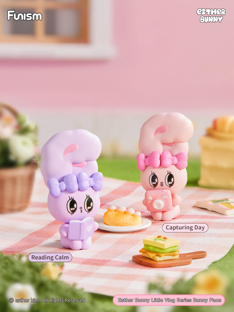 Picnic scene with one bunny reading calmly while another photographs the moment with a pink camera. They sit on a gingham blanket with food props like sandwiches and cake, evoking a cheerful outdoor lifestyle.