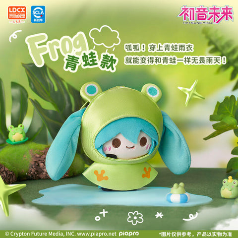 Frog-themed Hatsune Miku plush wearing a lime-green raincoat with big frog eyes on the hood and orange paw prints on the poncho. Her turquoise hair drapes around the outfit while she stands on a puddle. Background includes small frog figurines and leafy greenery, capturing a cute rainy forest atmosphere.