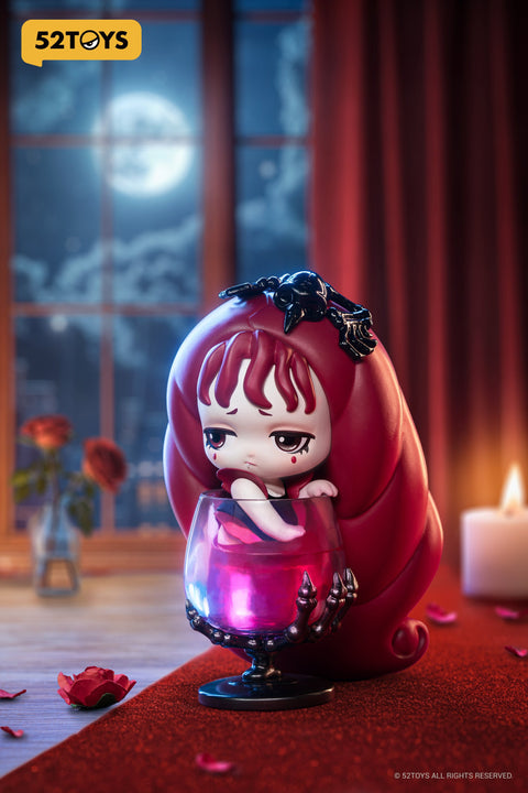 Close-up of the “Blood Chalice” figure from 52TOYS’ Lilith: The Century of Mary series. The character rests inside a translucent red goblet with a skeletal base, gazing somberly to the side. Her long red hair coils around the glass, adorned with a black ornament shaped like a bat or skull. The scene is set under moonlight, with red curtains, rose petals, and candles adding to the gothic, melancholic mood.