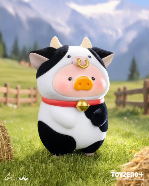 A LuLu the Piggy figure dressed as a black and white cow with small horns and a golden nose ring, wearing a red collar with a bell. The figure stands in a grassy farm scene with mountains and a wooden fence in the background.