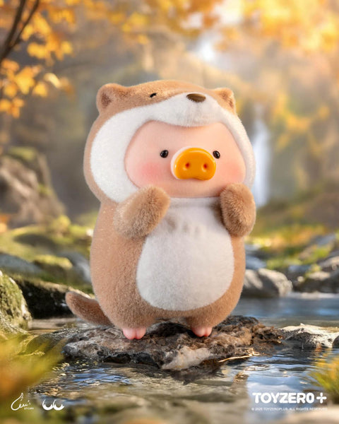 A LuLu the Piggy figure in a tan otter costume with a white belly, small rounded ears, and a tail. It stands on a river rock surrounded by clear water and forest greenery with autumn tones.