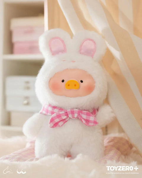 A soft and cuddly plush from the "LuLu The Piggy: Joyful Time Series" features LuLu dressed in a fluffy white bunny costume with long ears and a pink gingham bow. The plush has a round, blushing face with an adorable piggy snout, exuding warmth and charm. It sits on a cozy, pastel-colored bed with soft blankets, enhancing its comforting and huggable appeal. The background, featuring neutral tones and delicate decor, creates a serene and inviting atmosphere.