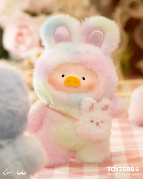 A delightful plush from the "LuLu The Piggy: Joyful Time Series" features LuLu dressed in a pastel rainbow bunny costume with soft, fluffy ears. It carries a tiny plush bunny bag, adding extra charm to its adorable design. With rosy cheeks and a gentle expression, the plush sits on a pink gingham tablecloth, surrounded by a warm, dreamy setting filled with soft flowers and blurred pastel hues, creating a cozy and whimsical atmosphere.