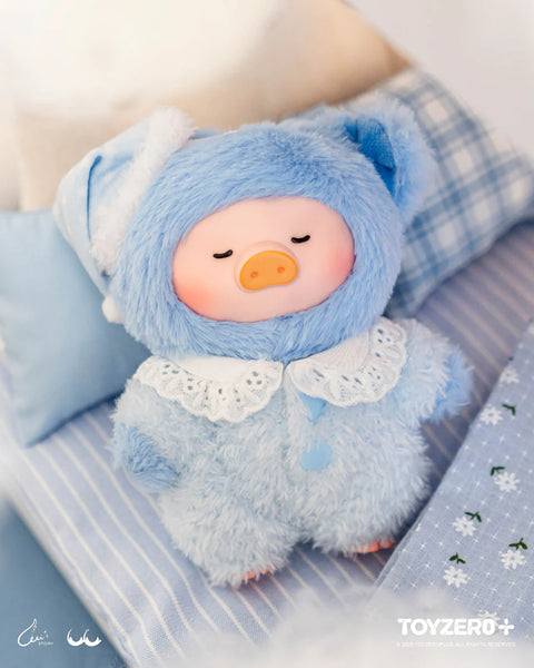 A blue plush pig dressed in a fuzzy bear suit sleeps peacefully on a striped bed. The suit features rounded ears, a lace collar, and blue buttons. A matching sleep hat with white trim rests on its head. The calm bedroom setting and closed eyes emphasize a sleep-well nighttime theme.