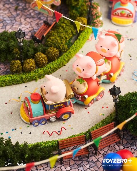 Three pig figurines ride in colorful carnival-themed mini train cars shaped like whimsical vehicles. Confetti is scattered along the curved path, with bunting overhead and hedges, benches, and streetlamps bordering the lively amusement park setting.