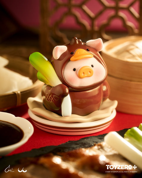 LuLu the Piggy wearing a glossy, brown roast duck costume holding green scallions. The figure sits on a white plate next to a dish of hoisin sauce and sliced duck meat.