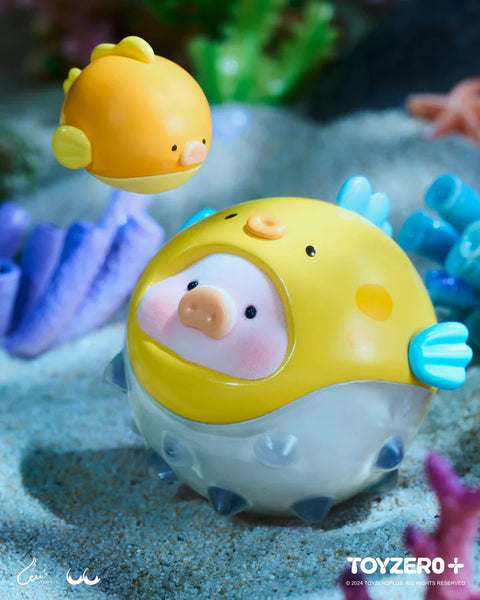 A LuLu the Piggy figure from the Ocean Series by 52TOYS and ToyZero+ is dressed as an adorable pufferfish. The pig’s face peeks out from a round, spiky costume with a bright yellow top and translucent lower half. A matching yellow fish companion floats nearby, both featuring pig-like snouts. Set against a sandy seabed with coral and sea plants, the scene captures a playful underwater encounter with a cute aquatic twist.