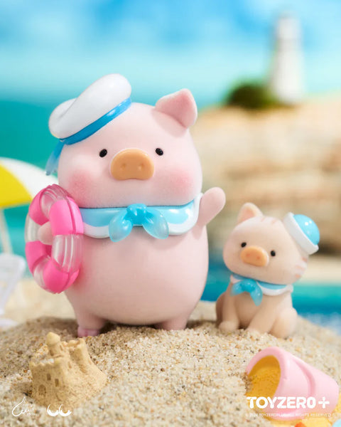 A LuLu the Piggy figure from the Ocean Series by 52TOYS and ToyZero+ stands on a sandy beach dressed as a sailor, wearing a white and blue sailor hat and scarf, and holding a pink float ring. Beside the pig is a cute cat companion in a matching outfit. A tiny sandcastle and overturned pail complete the scene, with a lighthouse and ocean in the background, evoking a cheerful and nostalgic day by the sea.