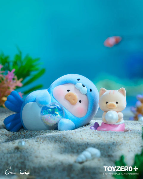 A LuLu the Piggy figure from the Ocean Series by 52TOYS and ToyZero+ is dressed in a soft blue seal costume, lying on the sandy ocean floor while holding a sparkling orb. Beside it sits a small cat companion with a mermaid tail, clutching a pearl. Surrounded by seashells, plants, and underwater life, the scene radiates a peaceful, magical deep-sea vibe with a pastel color palette and gentle expressions.