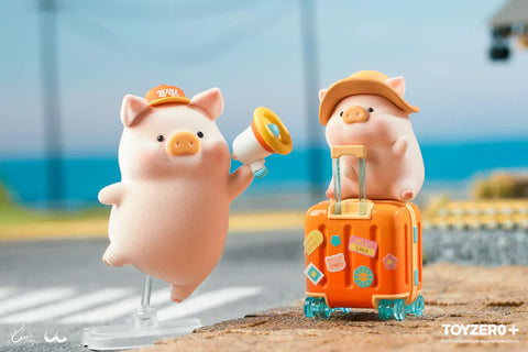 Two LuLu the Piggy figurines from the Travel Series by 52TOYS and ToyZero+ are featured in a seaside setting. One pig, wearing an orange cap, floats mid-air while holding a megaphone. The other, in a wide-brimmed hat, sits on a bright orange suitcase decorated with travel-themed stickers. Both figures capture a playful and adventurous vacation vibe, with a scenic coastal backdrop and train tracks in the distance.