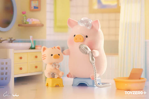 A close-up of a scene from the “Lulu the Piggy: Stay With You” blind box series shows a pink piggy character standing on a blue stool in a pastel-colored bathroom. With bubbles on its head, the piggy holds a silver showerhead, preparing for a wash. Beside it, a small striped cat figure stands on a yellow stool with shampoo bubbles on its head, mimicking the pig’s pose. The background features soft yellow and blue tones, a sink, a mirror, and a laundry basket, creating a warm, playful atmosphere.