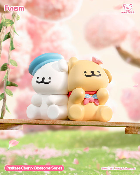 A white Maltese bear with a blue cap and a tan Maltese bear with a pink bow lean gently against each other while seated on a swing. Behind them, pink cherry blossoms create a soft, romantic springtime backdrop.