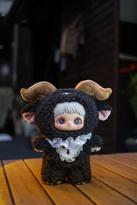 Black curly plush doll featuring brown ram horns, pink inner ears, and a white ruffled Victorian-style collar with a cross detail. Its eyes have a plus and minus symbol.