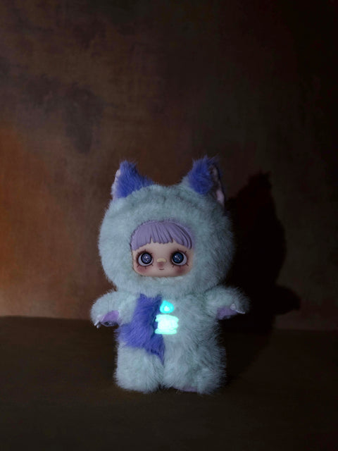 Low-light shot of the Ghost Elda doll showing a glowing turquoise candle icon on its chest. The plush fur appears light mint green in the dark.