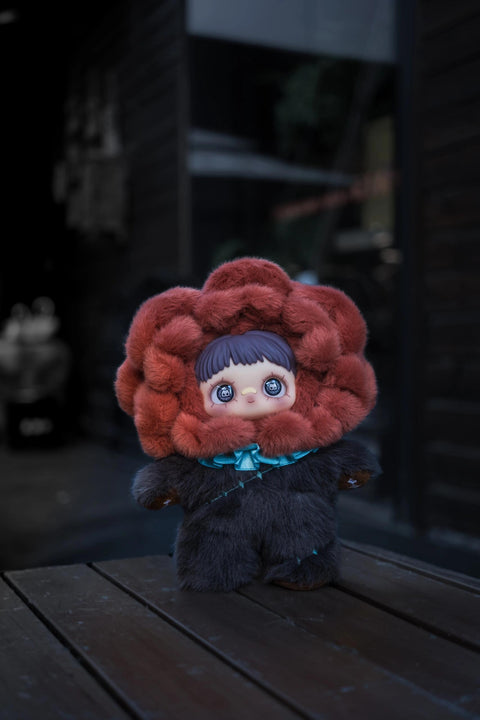 Plush doll with a dark grey body and a large, deep red ruffled collar resembling a rose around its head. It has blue eyes with skull patterns and a small turquoise bow at the neck.