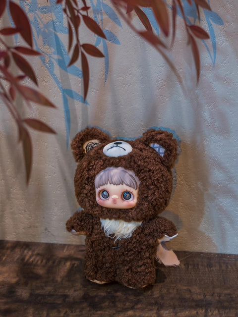 Brown textured plush bear doll with a sad-faced white patch on its head and plaid fabric inside one ear. It has blue-toned eyes and a white tuft of fur on its chest.