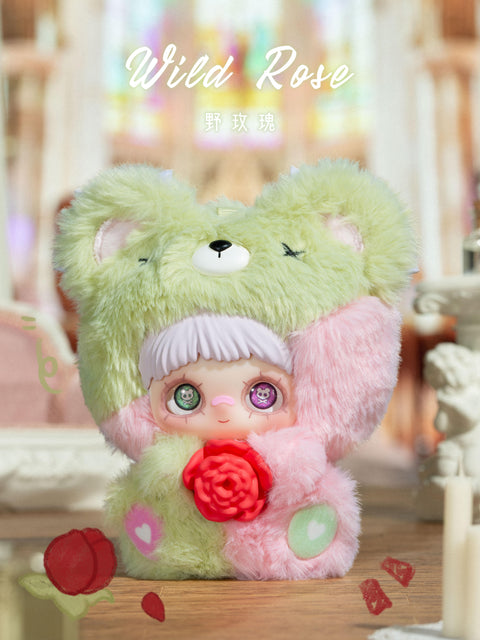 A plush figure stands on a wooden surface wearing a fuzzy hood split between light green and pale pink, with rounded ears and a small cream nose detail on top. The character has white bangs, large patterned eyes, and holds a red rose at the center. The background features soft pastel architecture, candles, and scattered rose petals under warm lighting.