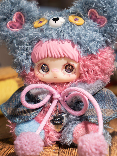 A small plush figure sits upright wearing a fuzzy light-blue and gray bear hood with yellow button eyes and heart-shaped ear patches. The character has short gray bangs, large printed eyes, and a soft cape tied with pale pom-pom cords. The body features matching plush textures in muted blue tones.
