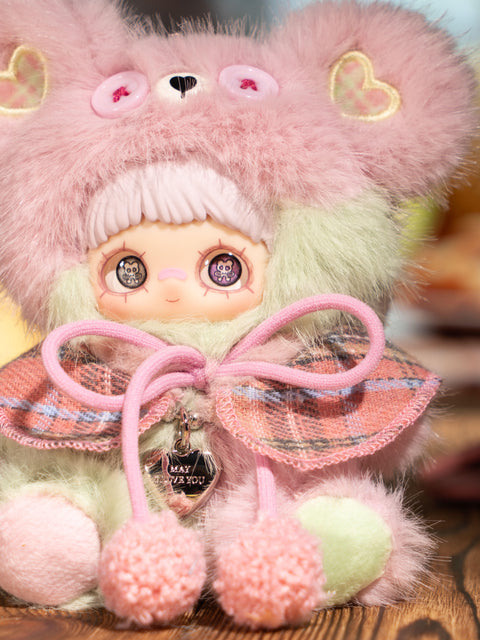 A pastel plush figure wears a pale pink and mint fuzzy bear hood with pink button eyes and heart ear patches. The character has light bangs, printed eyes, and a soft plaid cape tied with pink pom-pom cords. The body combines pastel green and pink plush textures in a candy-like color palette.