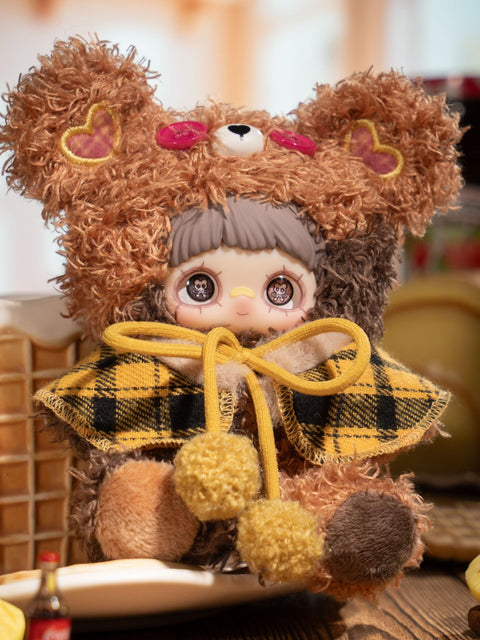 A plush figure sits wearing a brown fuzzy bear hood with red button eyes and stitched heart details on the ears. The character’s short dark bangs frame printed eyes. A red-and-brown plaid cape is tied at the front with pom-pom cords, while cookie stacks and warm-toned props fill the background.