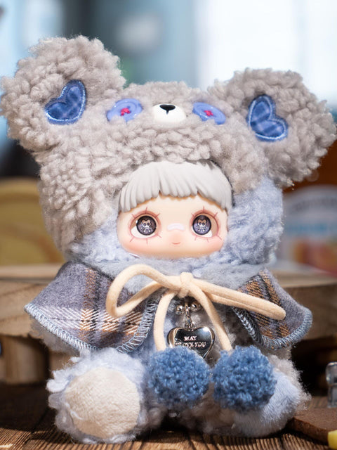 A plush figure sits upright wearing a soft gray fuzzy bear hood with blue button eyes and heart ear patches. The character has light gray bangs and printed eyes. A gray-blue plaid cape is tied with pale pom-pom cords, and the plush body features cool-toned blue and gray textures.