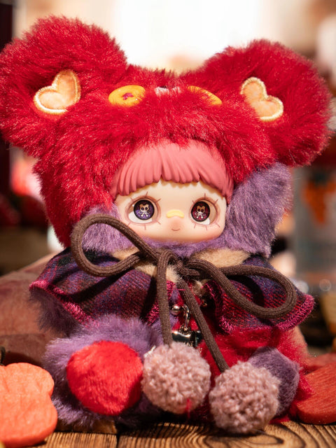 A hidden chaser plush figure wears a dark fuzzy bear hood with purple button eyes and heart-shaped ear patches. The character has pink bangs and printed eyes. A muted plaid cape is tied with pom-pom cords, and the plush body blends deep berry, brown, and pink tones among dessert props.