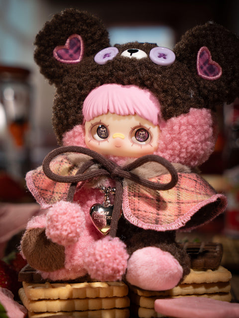 A plush figure sits on stacked cookies wearing a dark brown fuzzy bear hood with lavender button eyes and heart ear patches. The character has pink bangs and printed eyes. A pink-toned plaid cape is tied with pom-pom cords, and the plush body mixes brown and soft pink textures.