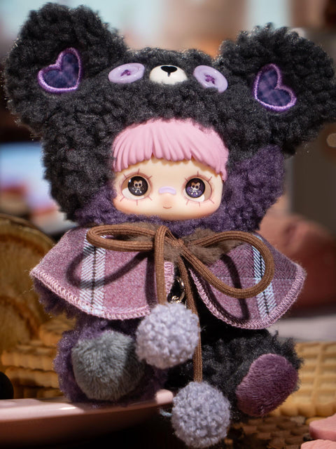 A plush character wears a dark gray fuzzy bear hood with lavender button eyes and purple heart ear patches. The figure has pink bangs, printed eyes, and a plaid cape tied with gray pom-pom cords. The plush body combines charcoal, lavender, and muted purple textures in a dessert-inspired palette.