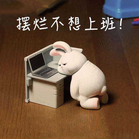A moody bunny figurine in a blue tie slumps against a desk with a laptop, glaring at the screen, humorously embodying work frustration and goblin-mode apathy.