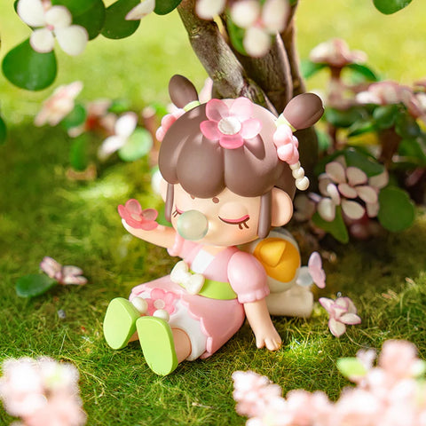 A peaceful collectible figurine from the "Nanci Blooming Girl" series features a girl dressed in a pink and white outfit, sitting under a blossoming tree. Her twin buns are adorned with delicate flower ornaments, and she holds a pink flower in her hand while blowing a bubblegum bubble. With closed eyes and a serene expression, she enjoys the gentle fall of petals around her. The lush green grass and vibrant blossoms create a tranquil and dreamy atmosphere, capturing a moment of pure relaxation.