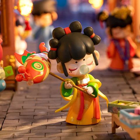 A festive collectible figurine from the "Nanci Blooming Girl" series features a girl dressed in a vibrant yellow and green traditional outfit, holding a colorful fish-shaped lantern on a stick. Her black hair is styled in intricate buns with red accents, and she blows a bubblegum bubble while enjoying the lively market scene. Warm lantern lights illuminate the background, filled with bustling figures, capturing the joy and charm of a traditional festival celebration.