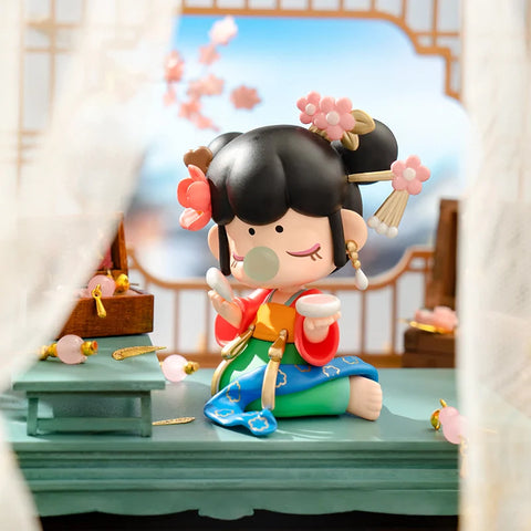 A charming collectible figurine from the "Nanci Blooming Girl" series features a girl dressed in a colorful traditional outfit, secretly applying makeup. She holds a small mirror while delicately dabbing powder on her face, with an expression of playful mischief. Her black hair is adorned with intricate floral hairpins and golden tassels. Set in an elegant room with a vanity table, jewelry, and soft curtains, the scene captures a nostalgic and whimsical moment of youthful curiosity.