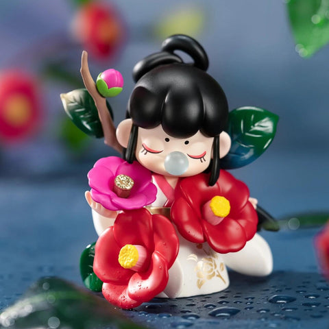 A figurine from the Nanci “Flower Stories” blind box series inspired by the camellia flower. The character has black hair styled in a top bun and wears a white outfit with gold floral details. She is surrounded by large red and pink camellia blossoms and green leaves, and holds a small flowering branch. With eyes closed and a bubble at her lips, she exudes calm and graceful beauty against a backdrop of glistening blue petals and raindrops.