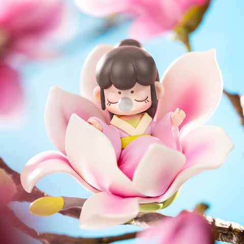 A figurine from the Nanci “Flower Stories” blind box series inspired by the magnolia flower. The character has black hair styled in a traditional bun and wears a pink and yellow hanfu-style outfit. Seated gracefully within a fully bloomed magnolia with soft white and pink petals, she blows a bubble with her eyes closed, surrounded by bright blue skies and blooming branches that evoke the elegance and beauty of spring.