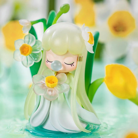 A figurine from the Nanci “Flower Stories” blind box series representing the narcissus flower. The character has soft pale green hair and wears a flowing white and green dress that mimics flower petals. She holds a delicate, translucent narcissus bloom and has a matching flower in her hair. With closed eyes and a bubble at her lips, she stands in a serene garden filled with blooming daffodils, radiating freshness and springtime charm.