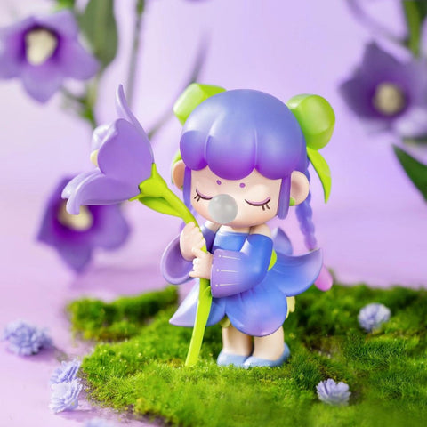 A figurine from the Nanci “Flower Stories” blind box series inspired by the platycodon flower. The character has purple hair styled in twin buns with green bows, and wears a petal-shaped purple and blue dress. She holds a large, delicate platycodon bloom close, eyes closed and blowing a bubble. Surrounded by moss and soft lavender tones, she radiates quiet elegance and whimsical charm.