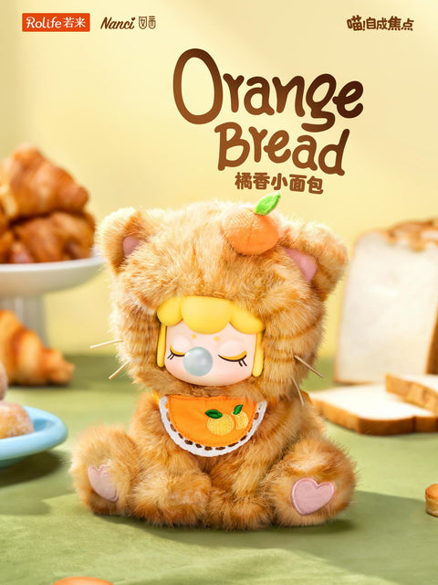 A bright ginger tabby plush Nanci with an orange fruit accessory on her head and an orange-patterned bib, surrounded by fresh bread and croissants.