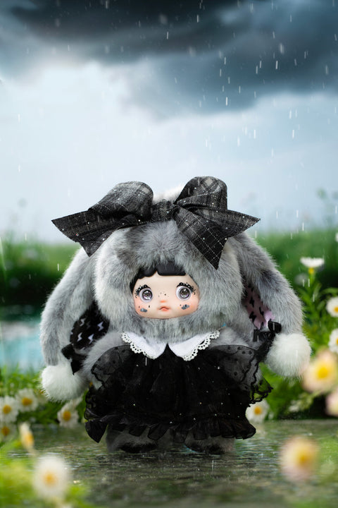 Plush character figure dressed in a dark gray hooded outfit with tall pointed ears and a metallic silver jacket, standing in shallow reflective water under stormy skies. The doll has large glossy eyes, subtle star markings on the cheeks, and thick fluffy fabric textures that emphasize a nighttime storm theme.
